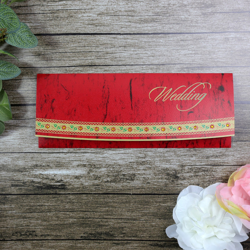 A red wedding invitation card with a decorative border and the word 'WEDDING' printed in gold lettering, placed on a wooden surface with a floral decoration to the side.